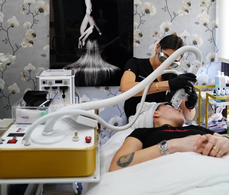 A technician performs a laser facial on a patient's face, who lies on a bed in a room with floral wallpaper.