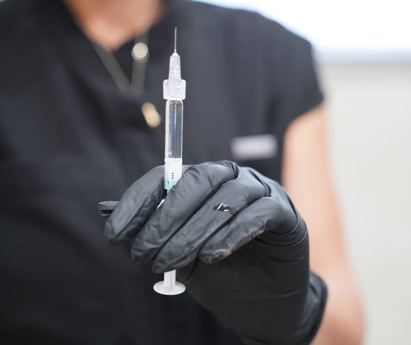 A gloved hand in a black scrub top holds a syringe with clear liquid and a needle.