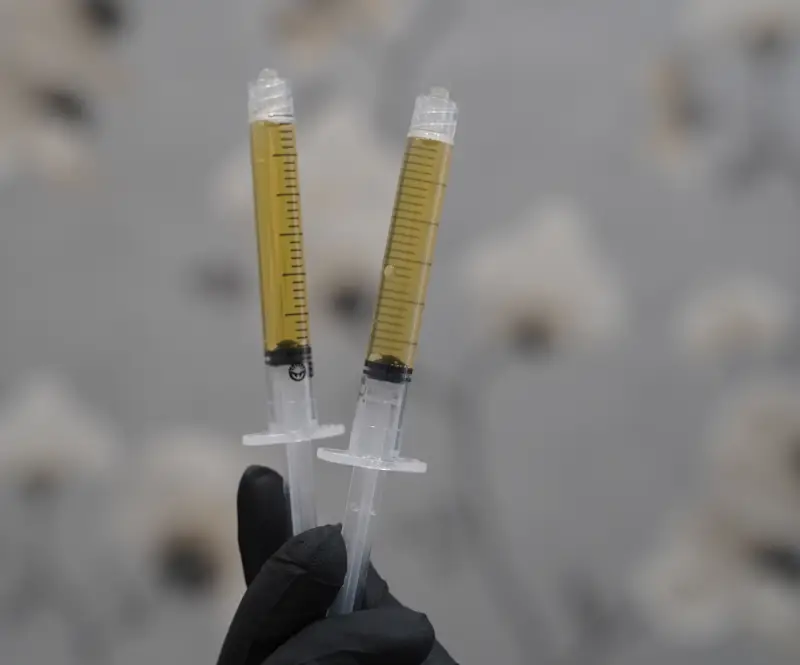A gloved hand in a black scrub top holds a syringe with clear liquid and a thin needle.