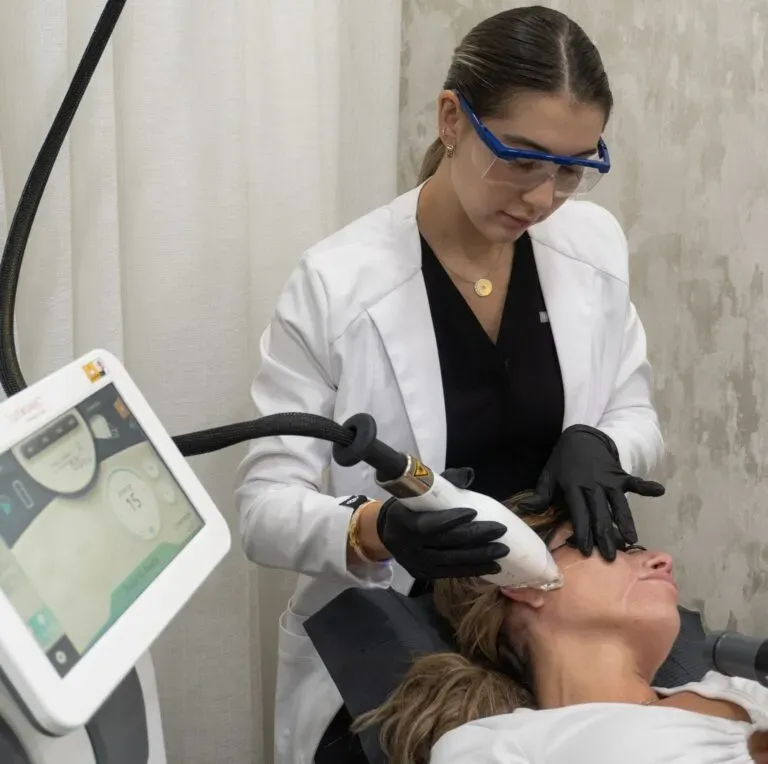 A person wearing safety glasses and lab coat performs a facial laser treatment on a reclining patient.