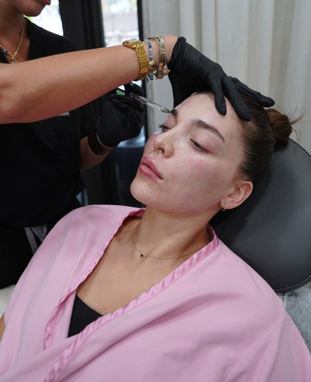A woman in a pink robe reclines as a healthcare professional injects solution near her eyebrow.