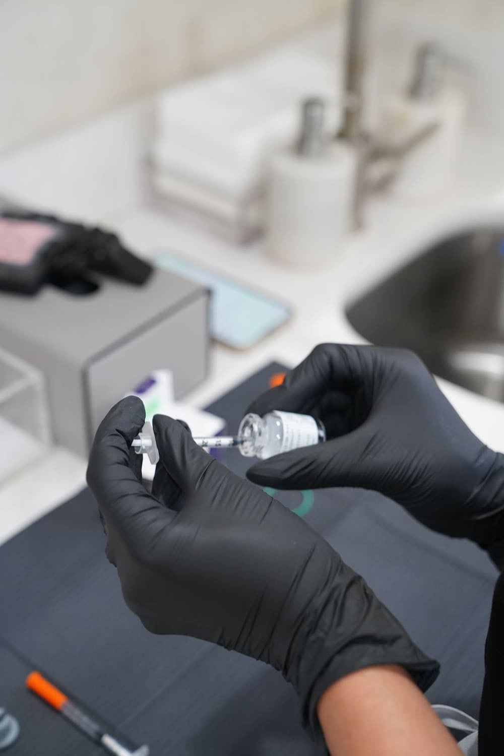 A person in black gloves draws clear liquid into a syringe from a small bottle on a dark mat.