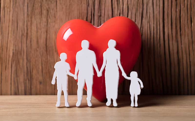 Four white paper figures of a family (two parents, two children) holding hands in front of a red heart.