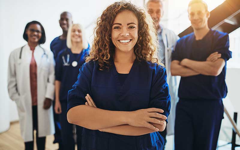 A smiling female medical professional with curly brown hair in blue scrubs, flanked by a diverse group of colleagues.