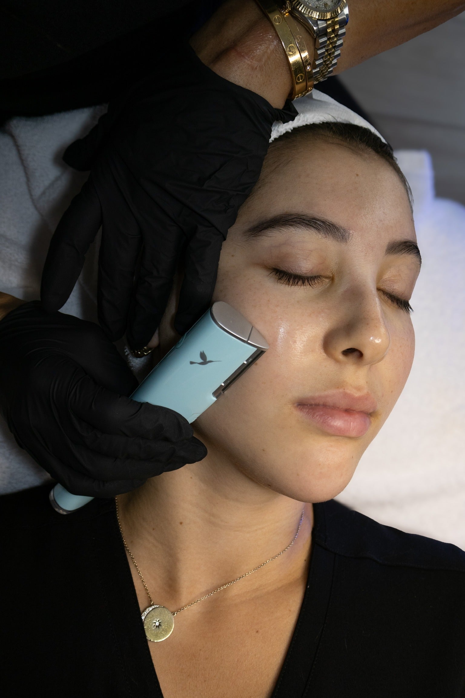 A person with eyes closed receiving a facial treatment with a light blue device from hands in black gloves.