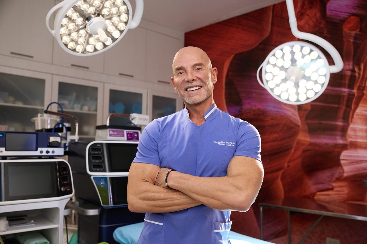 Smiling female beautician in a white lab coat sitting in a brightly lit cosmetology room with a magnifying lamp.