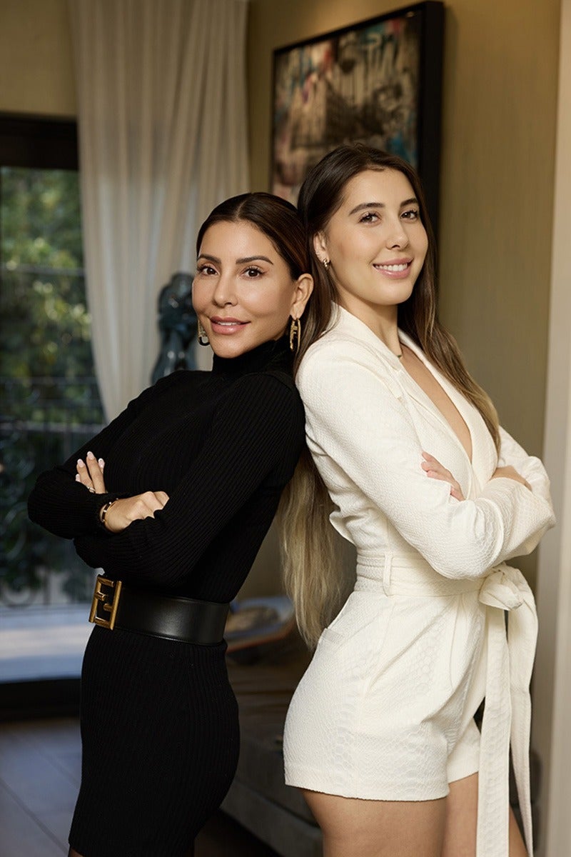 Two women, one in a fitted black ribbed dress and the other in a white romper, stand back-to-back, smiling.