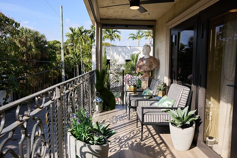 A sunny balcony with wrought iron railing, two striped chairs, potted plants, a bust, and palm trees in the background.