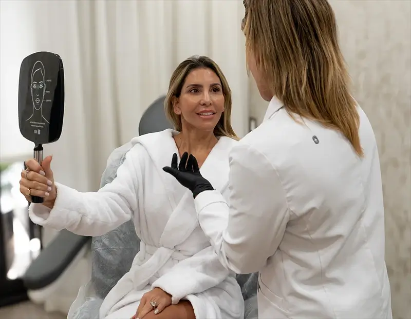 Woman in a robe holding a mirror with a face diagram, consulting with a medical professional in a white coat.