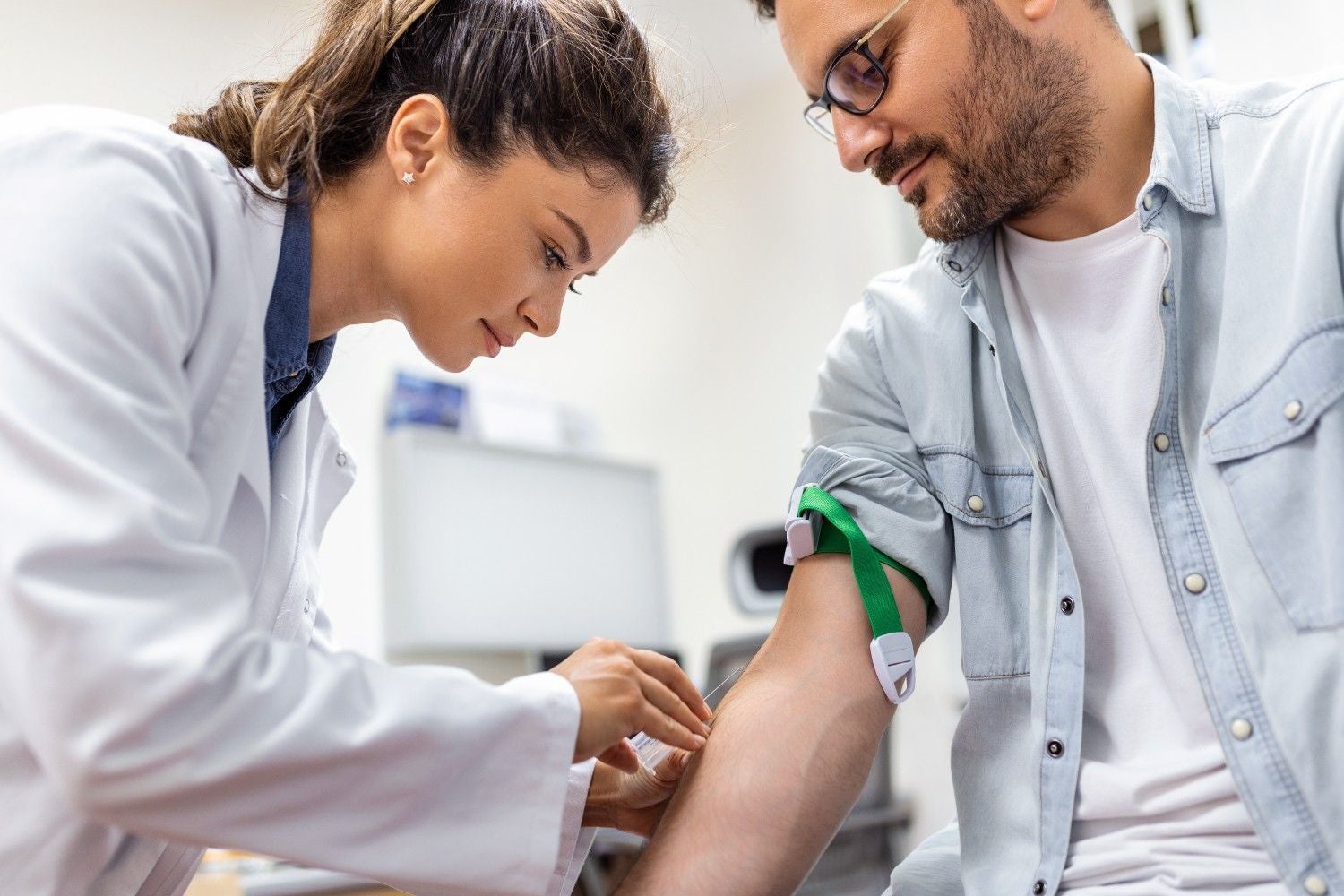 A phlebotomist in a white coat drawing blood from a male patient's arm, who wears a green tourniquet.