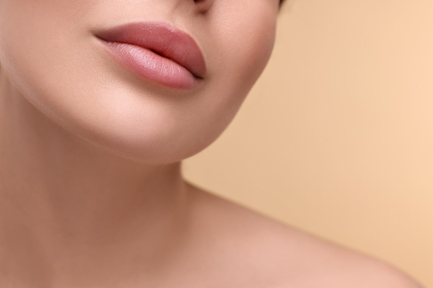 Lower left of a person's face showing glossy, plump pink lips and smooth skin against a cream background.